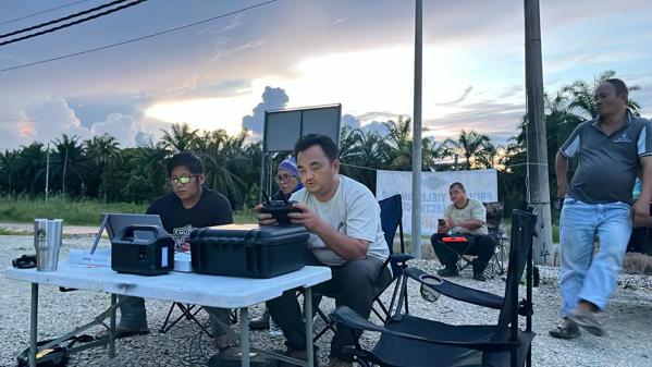 A photo showing Hutan researchers sitting in camp chairs and at collapsible tables. One is holding a controller for a drone. The sky is pale and edging toward dusk