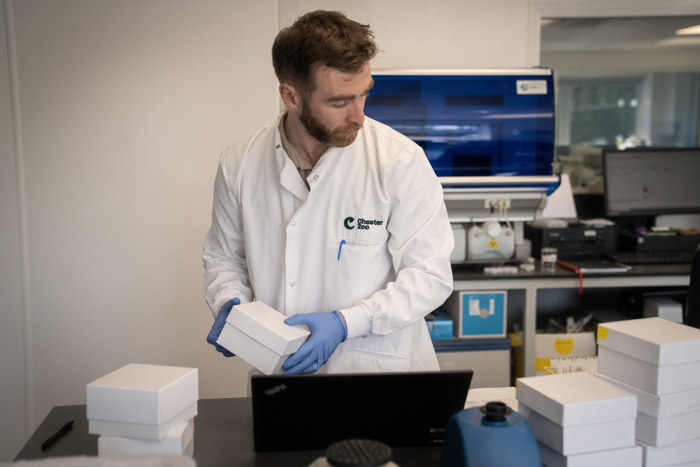 Photo shows a man in a lab coat moving boxes around a lab table