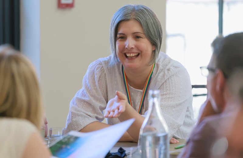 Charlotte Smith - a woman with chin-length grey hair and faintly freckled skin. She is smiling at the camera and is wearing a white shirt. She's chatting with people at a table
