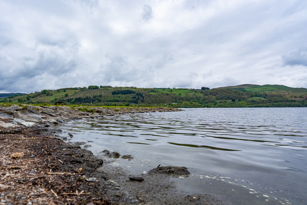 Photo shows Bala lake. The image is of a rocky shore and blue-grey water with rolling hills in the background
